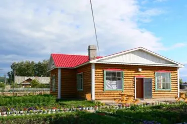 Shiwei Russian Ethnic Township,Hulunbuir,Chinatour Rustic log cabin with a red metal roof, green-trimmed windows, and a colorful flower garden in front.