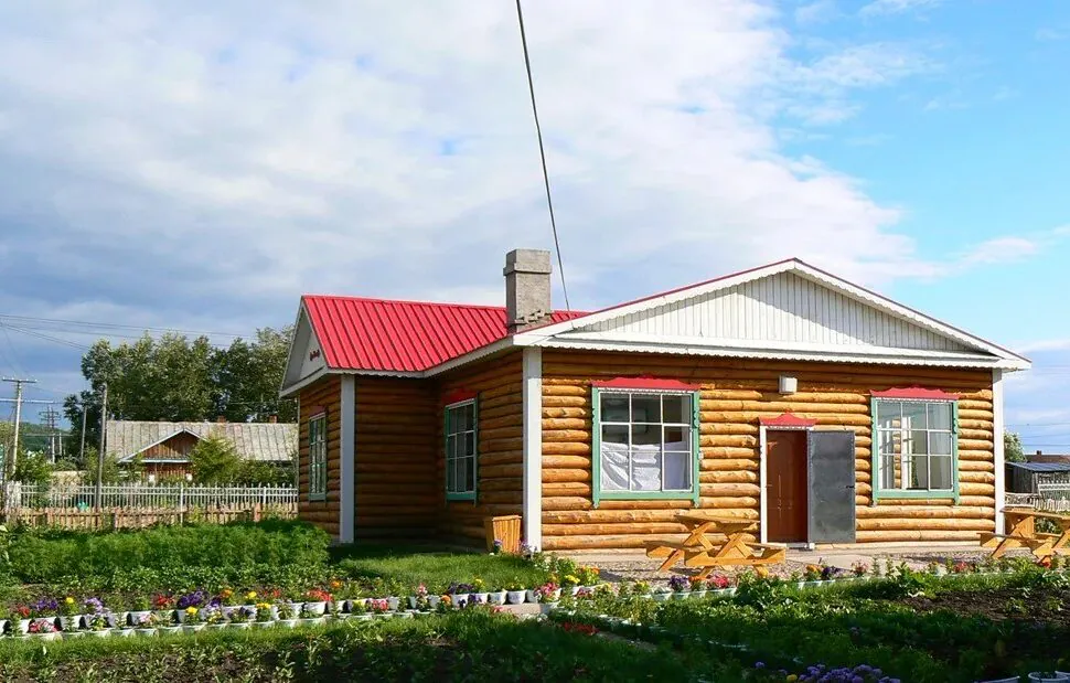 Shiwei Russian Ethnic Township,Hulunbuir,Chinatour Rustic log cabin with a red metal roof, green-trimmed windows, and a colorful flower garden in front.