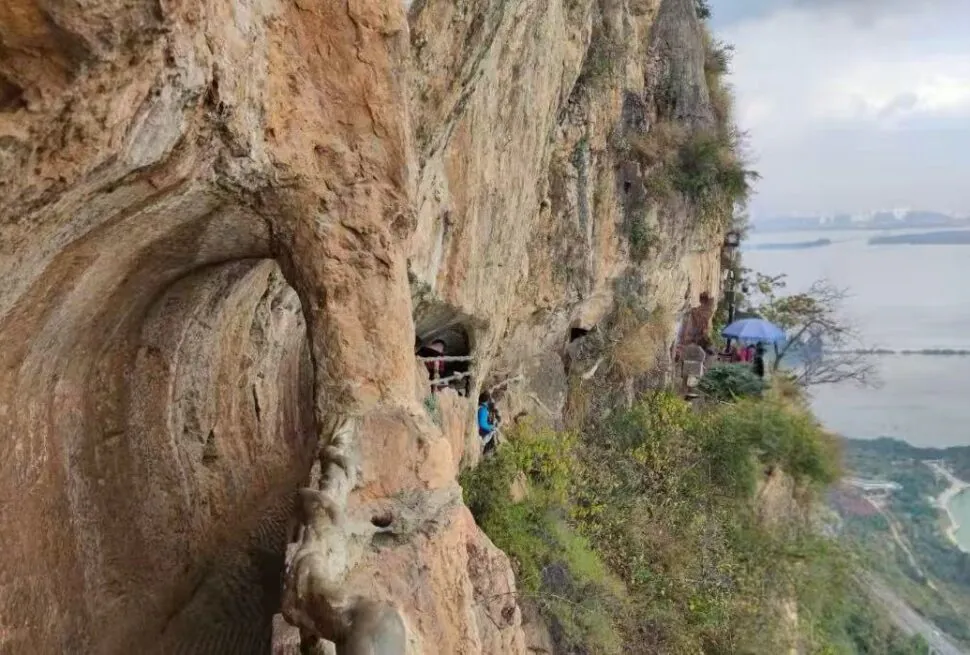 Hikers walk along a narrow rocky cliff path with carved railings, overlooking a large body of water in the distance.Western Hills & Dragon Gate,Kunming,Yunnan ,Chinatravel