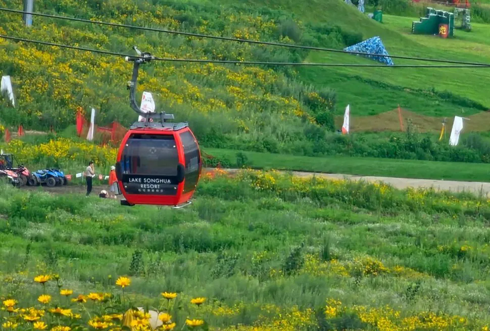 Vanke Lake Songhua Resort,Jilin,Chinatravel Red gondola cabin labeled Lake Songhua Resort, on a grassy hillside with yellow flowers nearby.