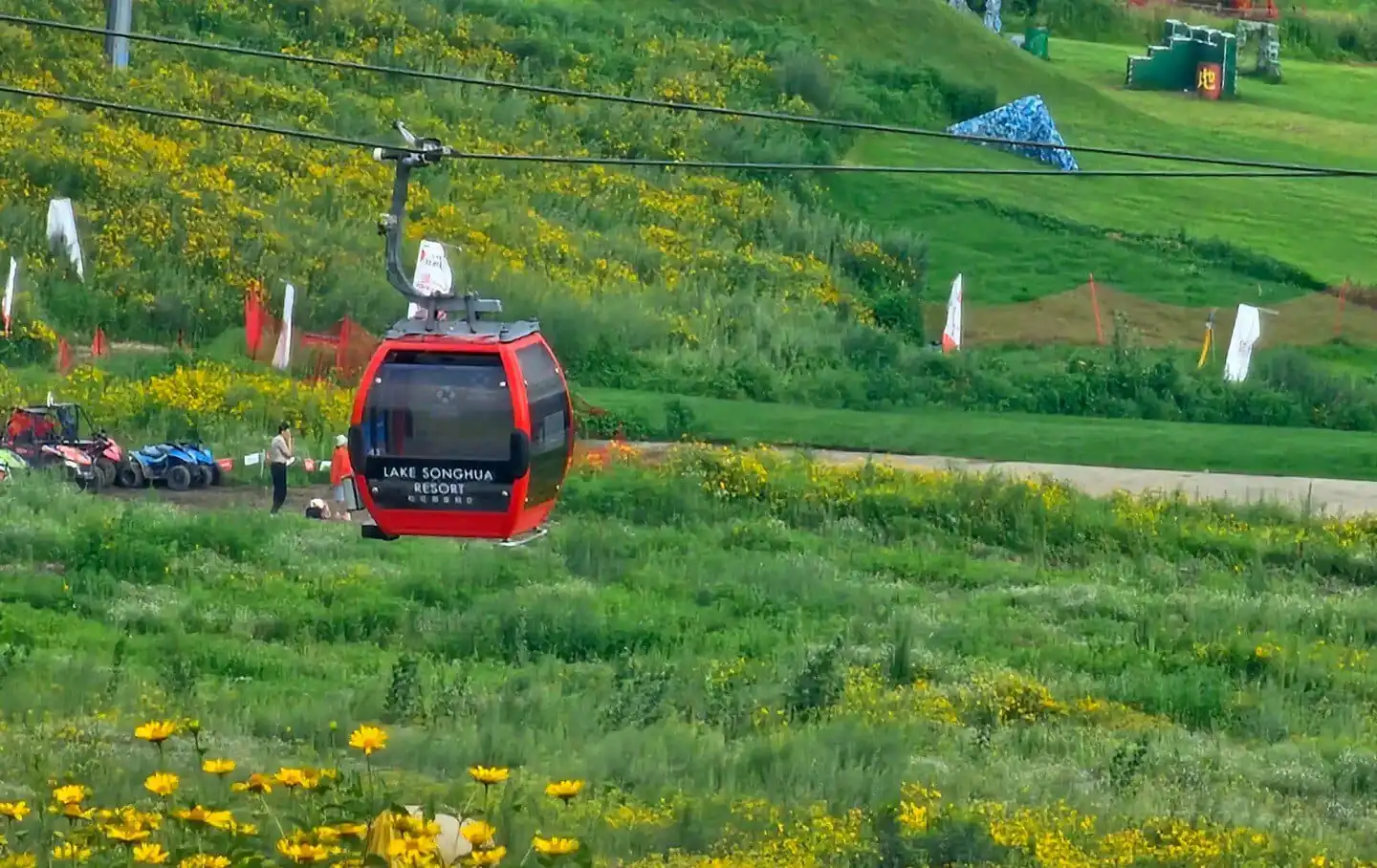 Vanke Lake Songhua Resort,Jilin,Chinatravel Red gondola cabin labeled Lake Songhua Resort, on a grassy hillside with yellow flowers nearby.