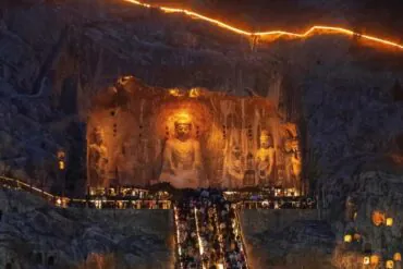 Longmen Grottoes,Luoyang,Chinatour Large Buddha statues carved into a cave wall with worshippers climbing illuminated stairs beneath them