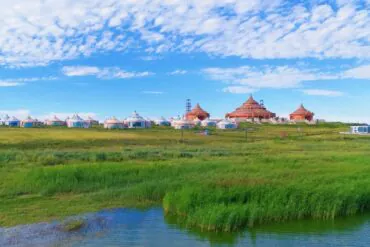 Row of white yurts and tan lodge-like buildings set along a grassy wetland under a bright blue sky and scattered clouds.Gegentala Grassland,Hohhot,Inner Mongolia,Chinatravel