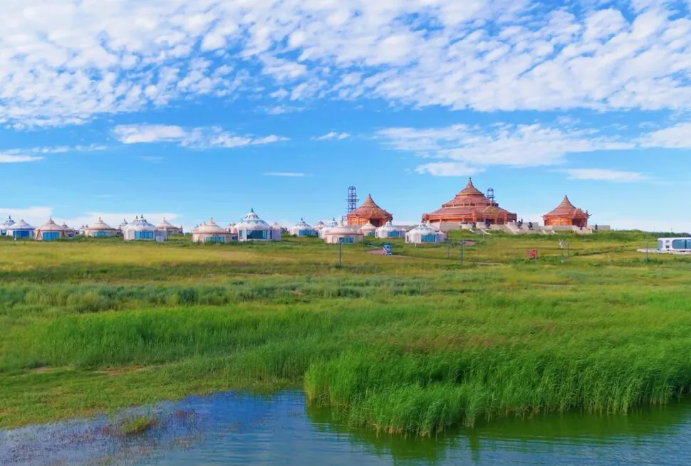 Row of white yurts and tan lodge-like buildings set along a grassy wetland under a bright blue sky and scattered clouds.Gegentala Grassland,Hohhot,Inner Mongolia,Chinatravel