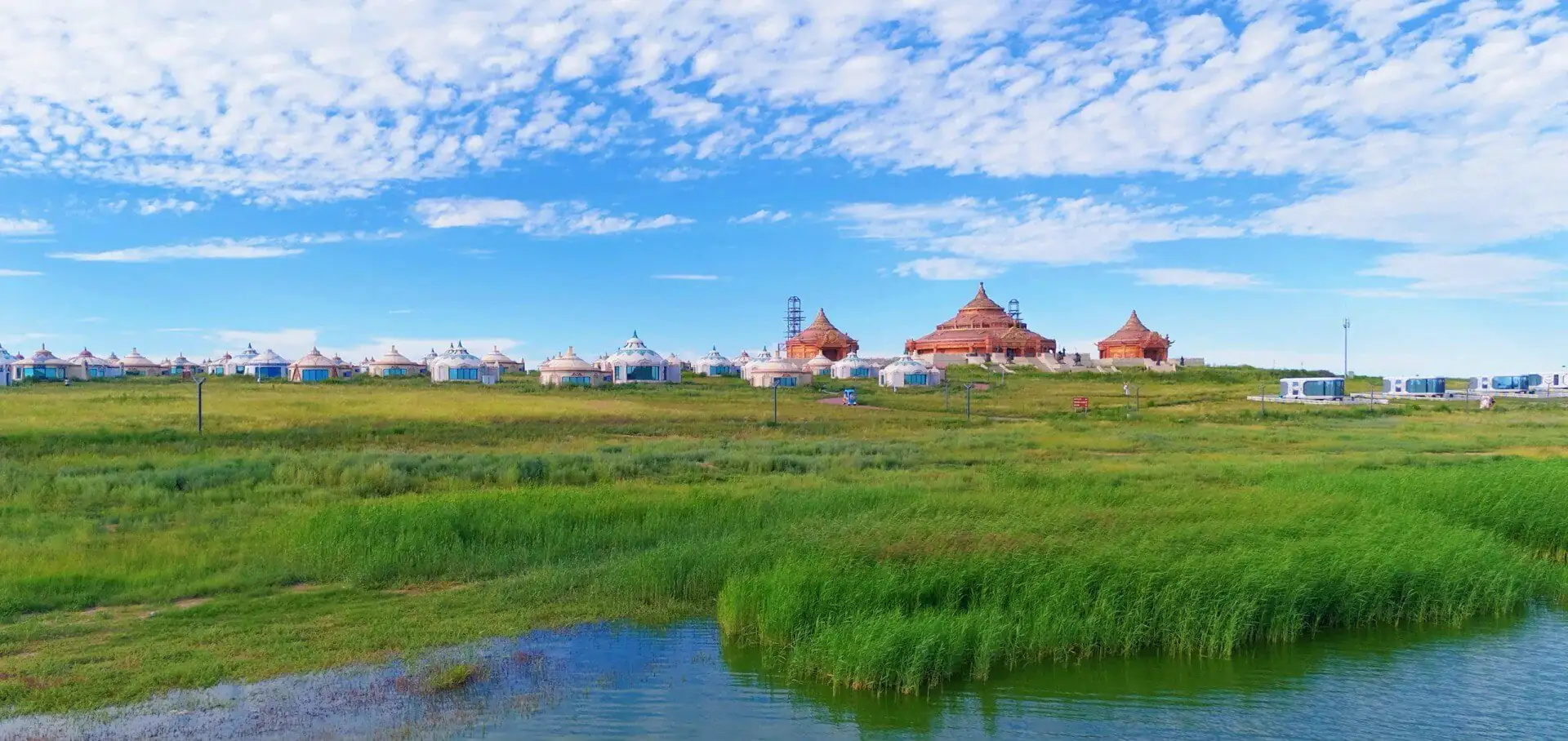 Row of white yurts and tan lodge-like buildings set along a grassy wetland under a bright blue sky and scattered clouds.Gegentala Grassland,Hohhot,Inner Mongolia,Chinatravel