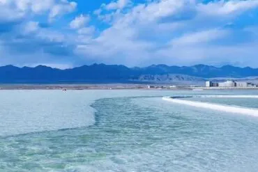 Turquoise shallow water with a curved white boardwalk leading to distant buildings and blue mountain range under a partly cloudy sky,Chaka Salt Lake,Xining,Qinghai