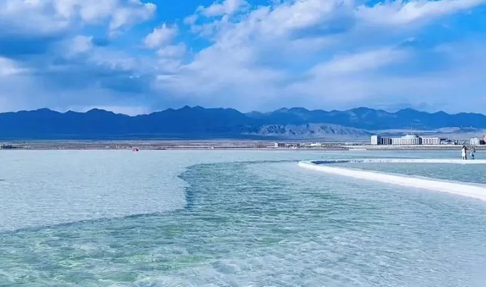 Turquoise shallow water with a curved white boardwalk leading to distant buildings and blue mountain range under a partly cloudy sky,Chaka Salt Lake,Xining,Qinghai