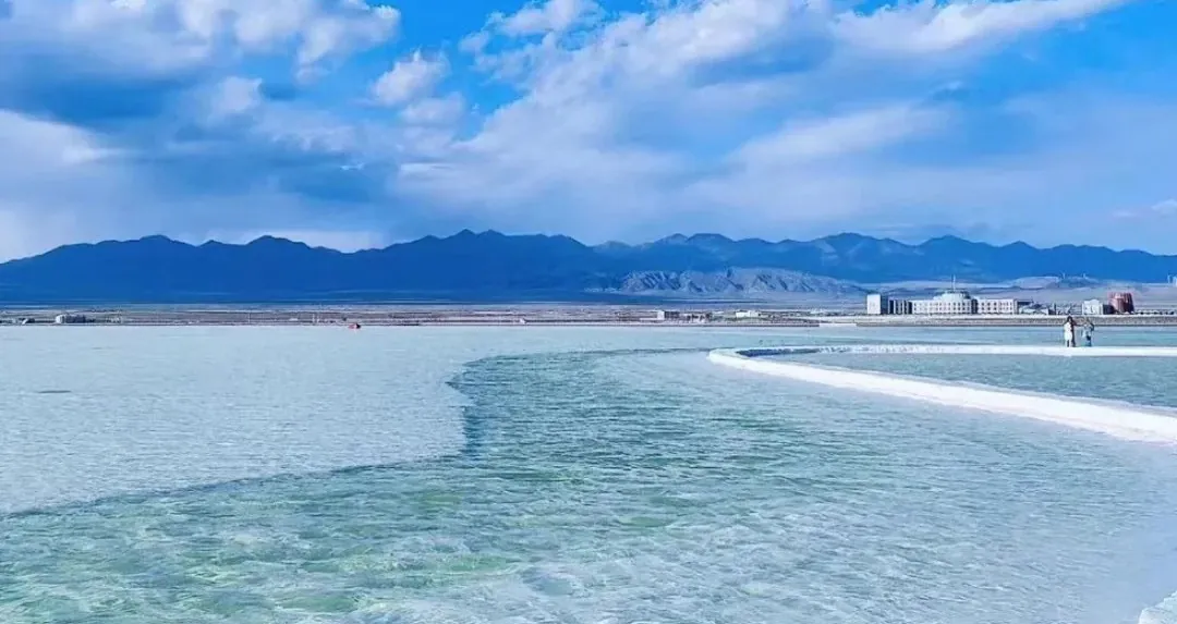 Turquoise shallow water with a curved white boardwalk leading to distant buildings and blue mountain range under a partly cloudy sky,Chaka Salt Lake,Xining,Qinghai