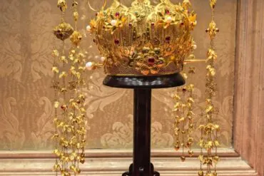 Ornate gold crown encrusted with red gems and pearls, displayed on a black pedestal with hanging gold chains around it.Guiyang White House,Guiyang,Guizhou