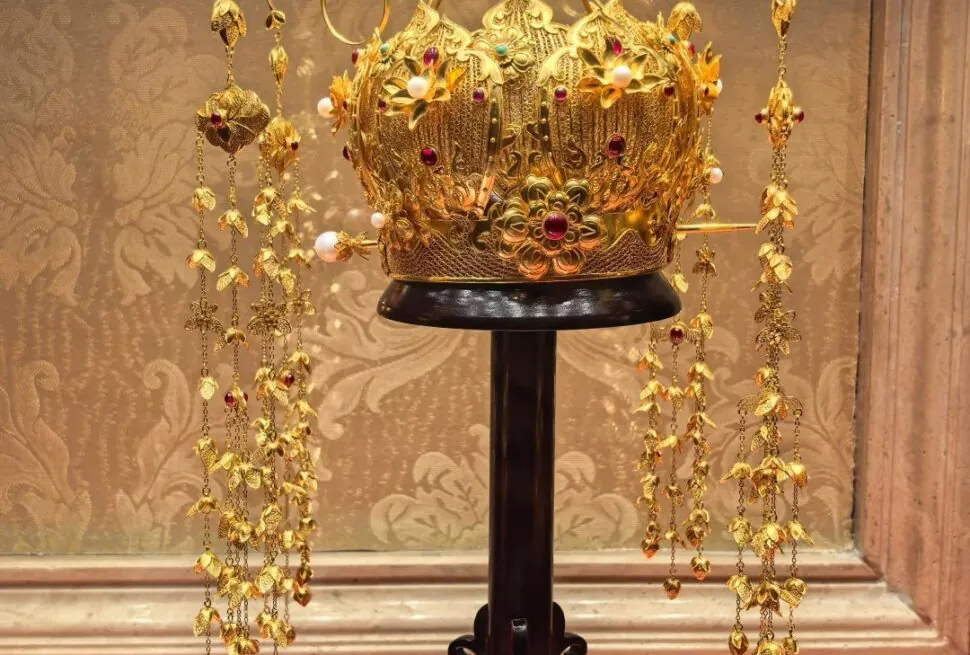 Ornate gold crown encrusted with red gems and pearls, displayed on a black pedestal with hanging gold chains around it.Guiyang White House,Guiyang,Guizhou