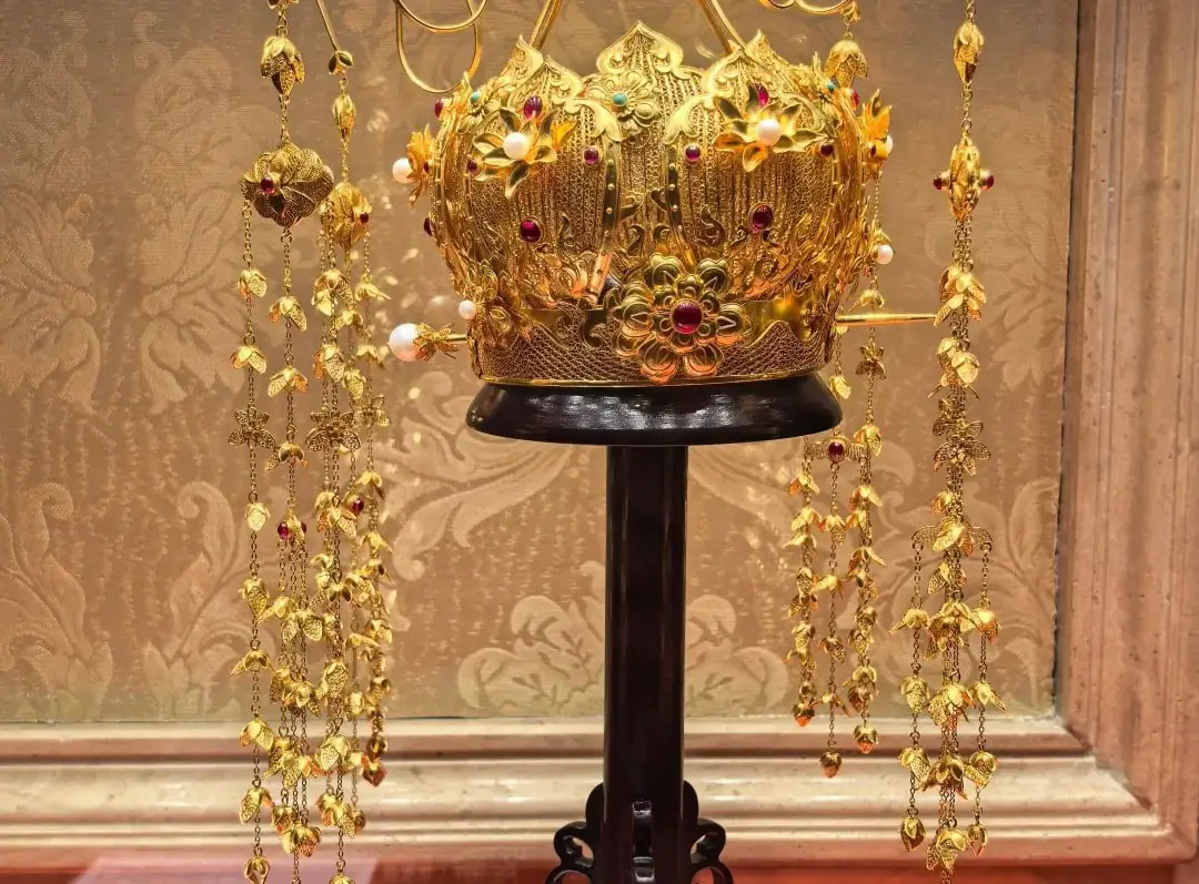 Ornate gold crown encrusted with red gems and pearls, displayed on a black pedestal with hanging gold chains around it.Guiyang White House,Guiyang,Guizhou