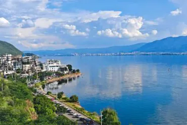 Lakeside town with white buildings along a rocky shore, green hills, and a calm blue lake with distant mountains under a cloudy sky.Erhai Lake,Dali,Yunnan,Chinatravel