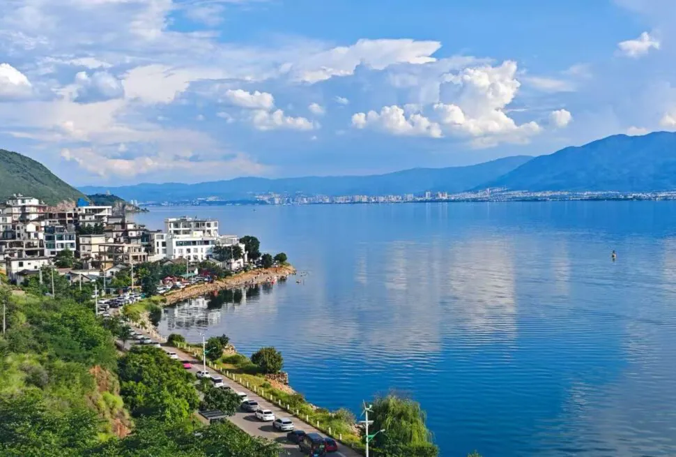 Lakeside town with white buildings along a rocky shore, green hills, and a calm blue lake with distant mountains under a cloudy sky.Erhai Lake,Dali,Yunnan,Chinatravel
