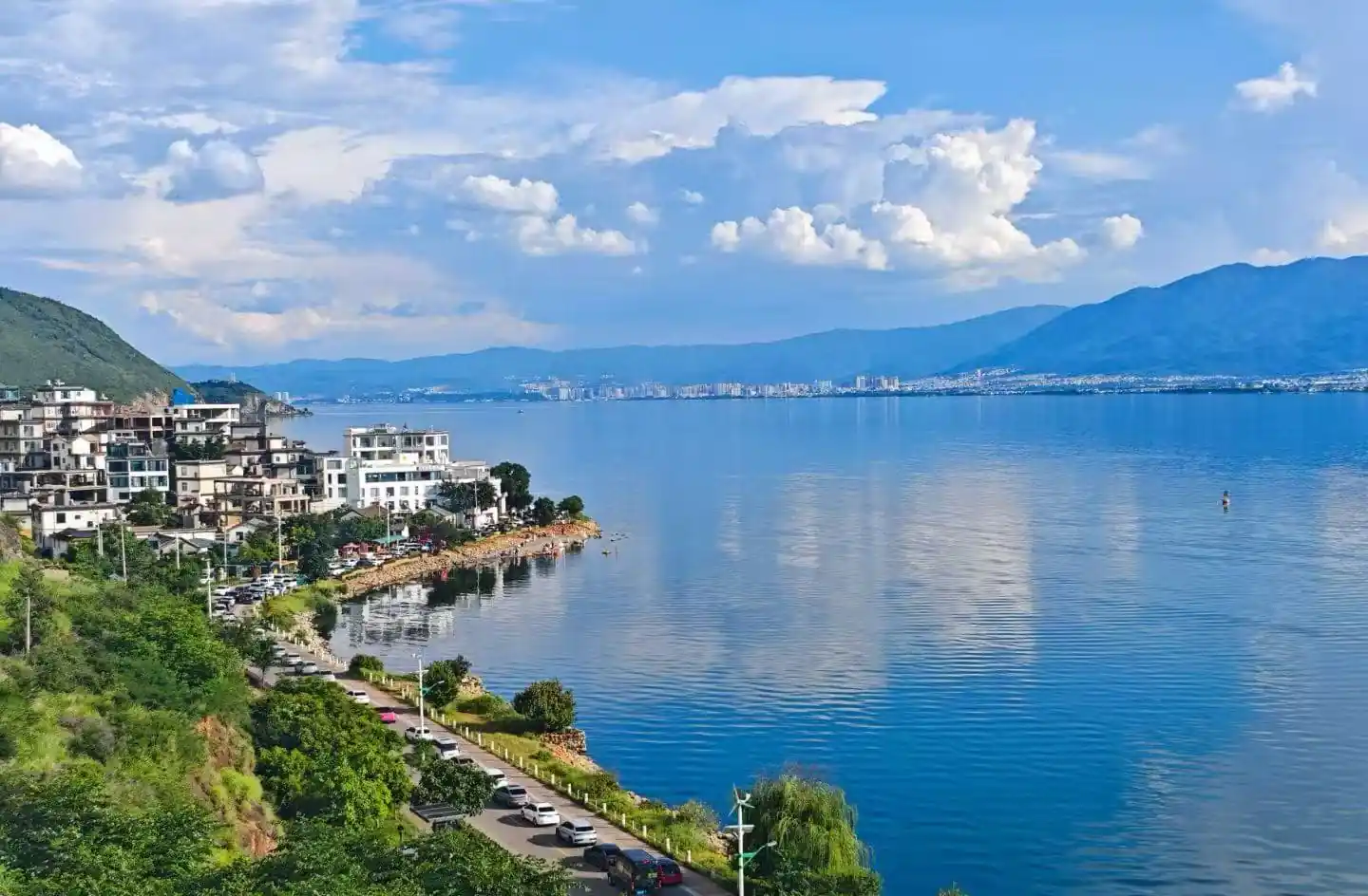 Lakeside town with white buildings along a rocky shore, green hills, and a calm blue lake with distant mountains under a cloudy sky.Erhai Lake,Dali,Yunnan,Chinatravel