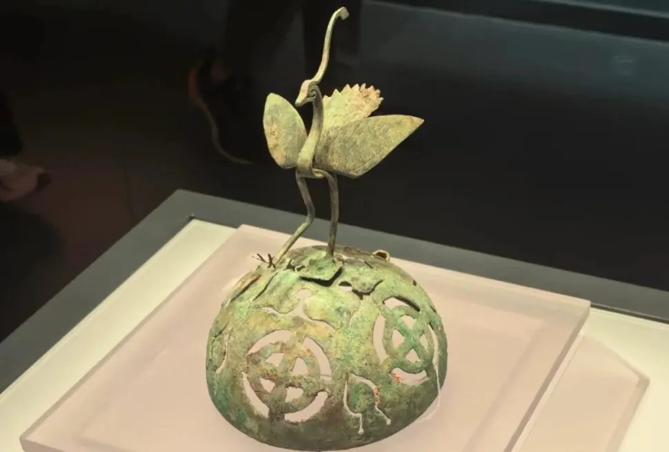 Bronze-green sculpture of two slender birds perched on a curved stem atop a perforated green sphere display piece in a museum case,Qinghai Provincial Museum,Xining,Qinghai