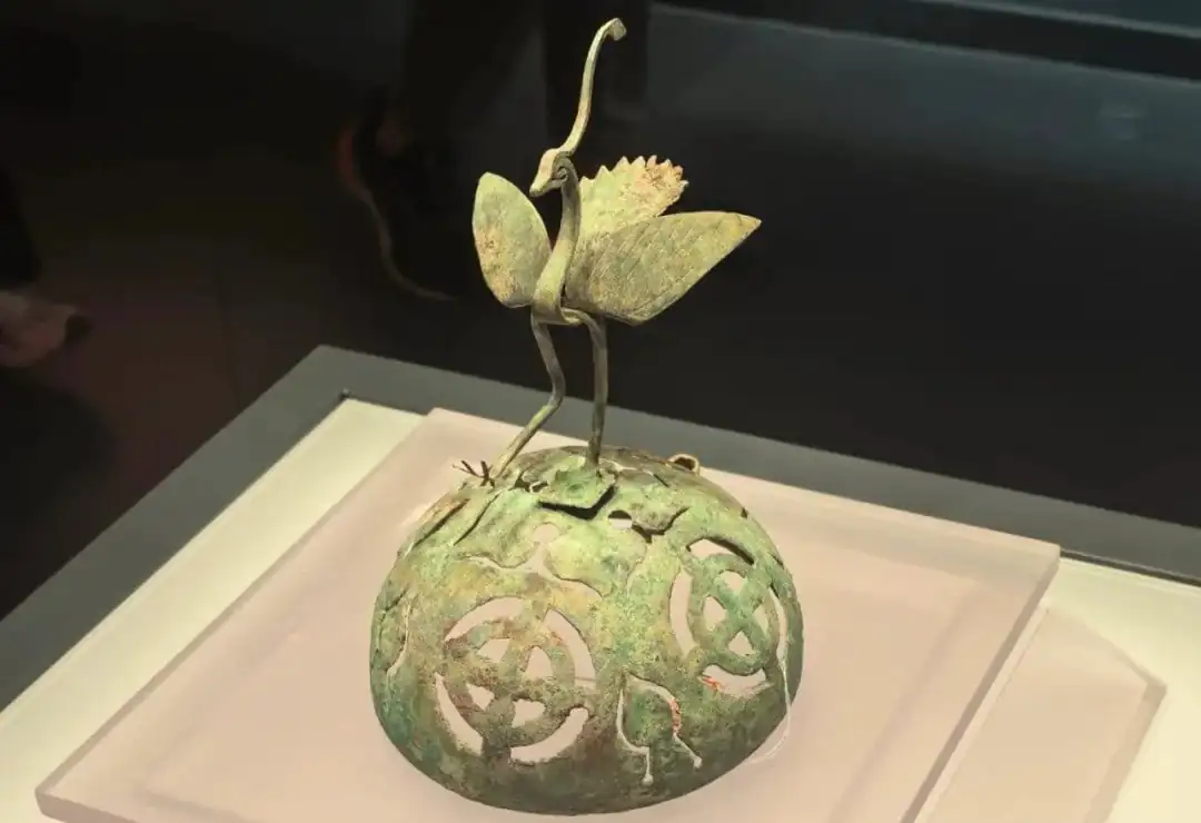 Bronze-green sculpture of two slender birds perched on a curved stem atop a perforated green sphere display piece in a museum case,Qinghai Provincial Museum,Xining,Qinghai