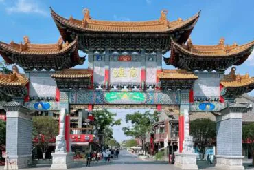 Ornate Chinese paifang archway with gold-tiled roofs and colorful carvings spans a street, flanked by shops and trees.Guandu Ancient Town,Kunming,Yunnan ,Chinatravel