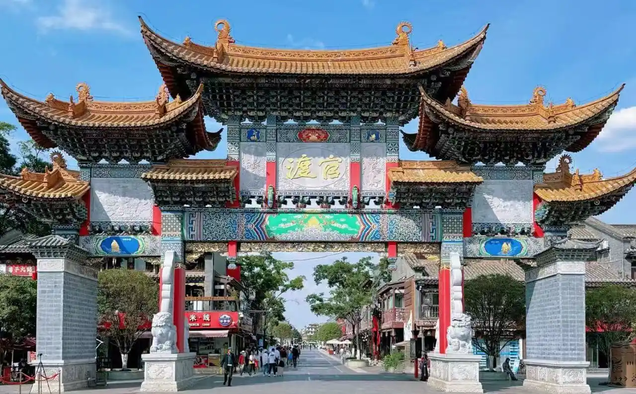 Ornate Chinese paifang archway with gold-tiled roofs and colorful carvings spans a street, flanked by shops and trees.Guandu Ancient Town,Kunming,Yunnan ,Chinatravel