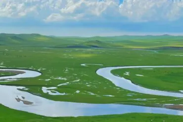 Mergel Gol River,Hulunbuir,Chinatour Wide green plain with a winding river curving through fields under a cloudy sky.