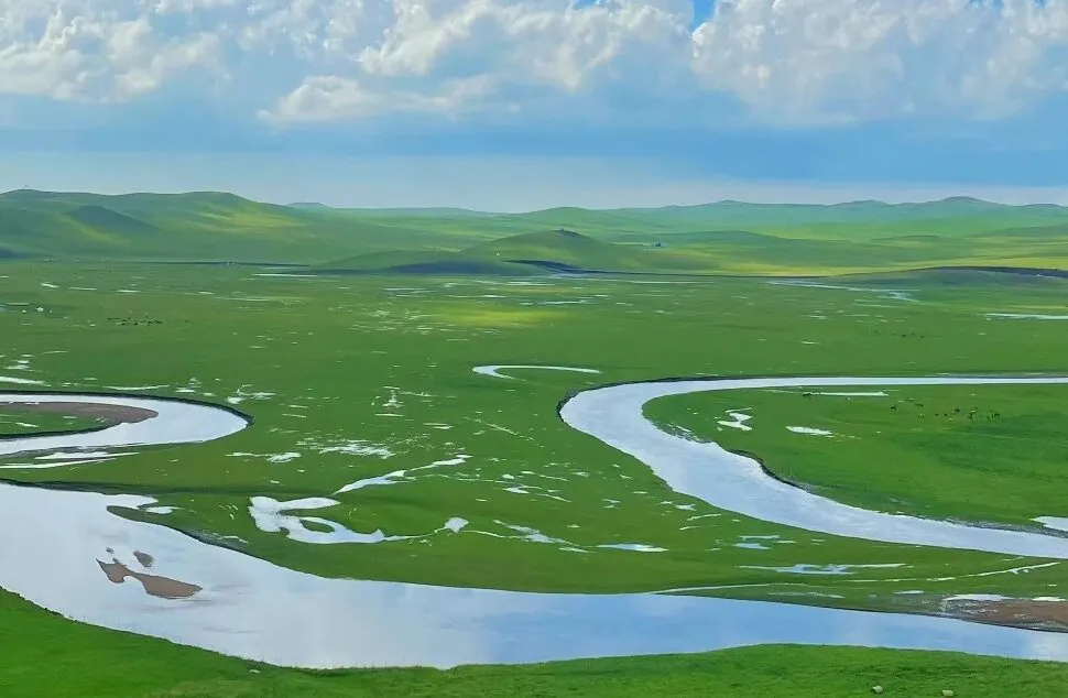Wide green plain with a winding river curving through fields under a cloudy sky.