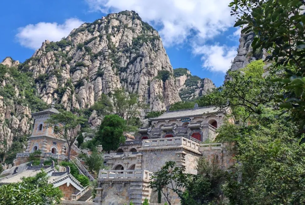 Chinese-style temple complex perched on a stone terrace against towering rocky cliffs, with red windows and carved stone railings amidst green trees.Sanhuangzhai,Shaolin & Songshan Area,Henan,Chinatravel