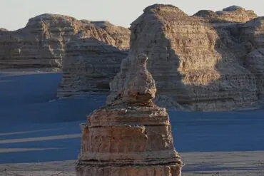 Layered sandstone formations in a desert with a prominent rock spire in the foreground and a fenced path at the base.Yardang National Geopark (Ghost City),Dunhuang,Gansu