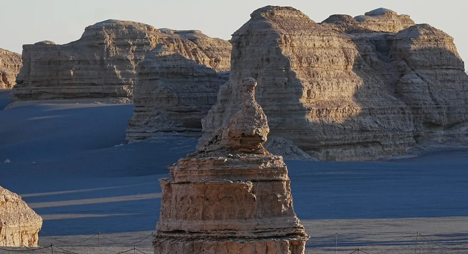 Layered sandstone formations in a desert with a prominent rock spire in the foreground and a fenced path at the base.Yardang National Geopark (Ghost City),Dunhuang,Gansu