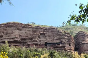 Rock cliff with carved cave temples and a wooden walkway along the base; a small traditional pavilion is built into the rock face.Tulou Temple Tibetan Buddhist Sites,Xining,Qinghai