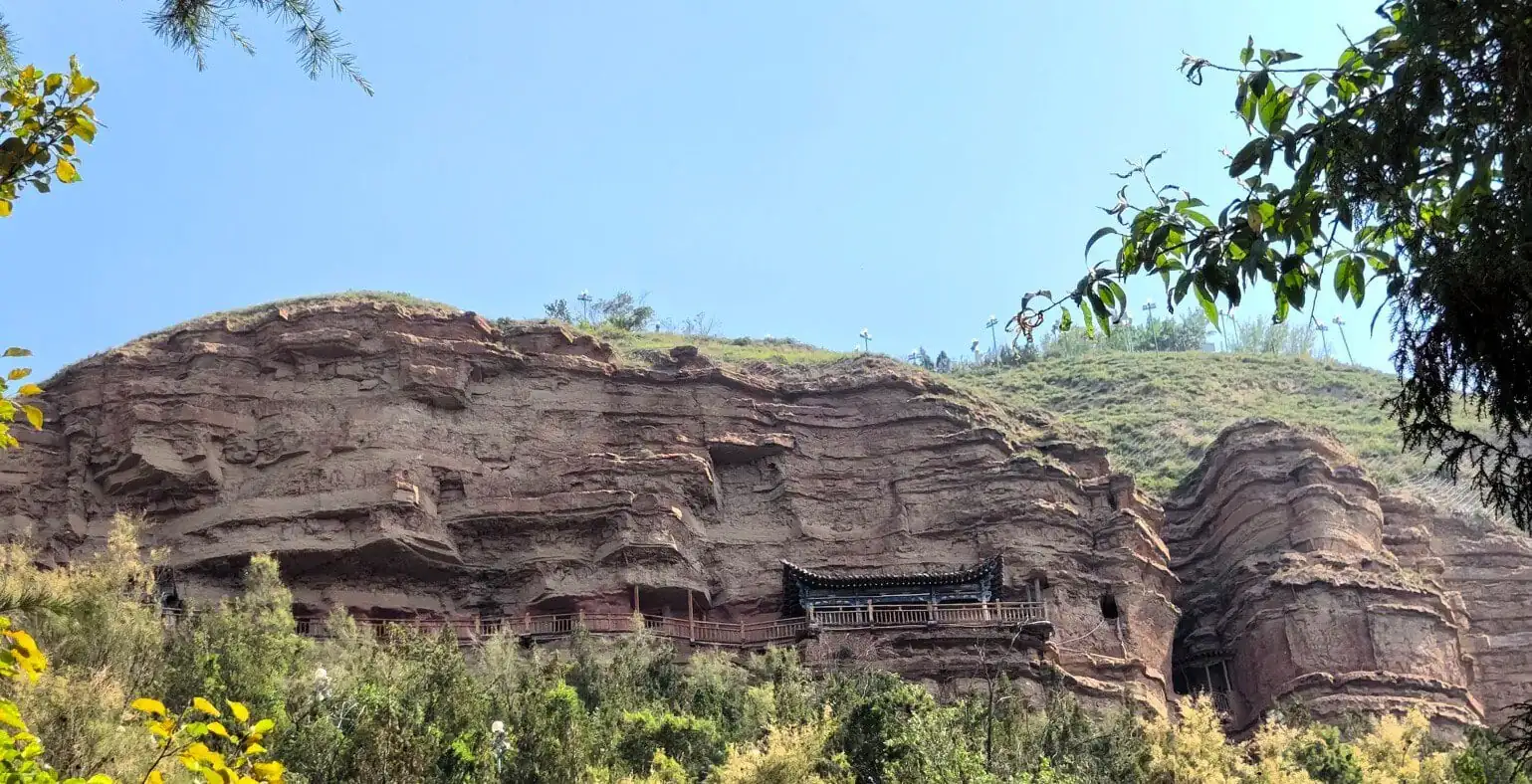 Rock cliff with carved cave temples and a wooden walkway along the base; a small traditional pavilion is built into the rock face.Tulou Temple Tibetan Buddhist Sites,Xining,Qinghai