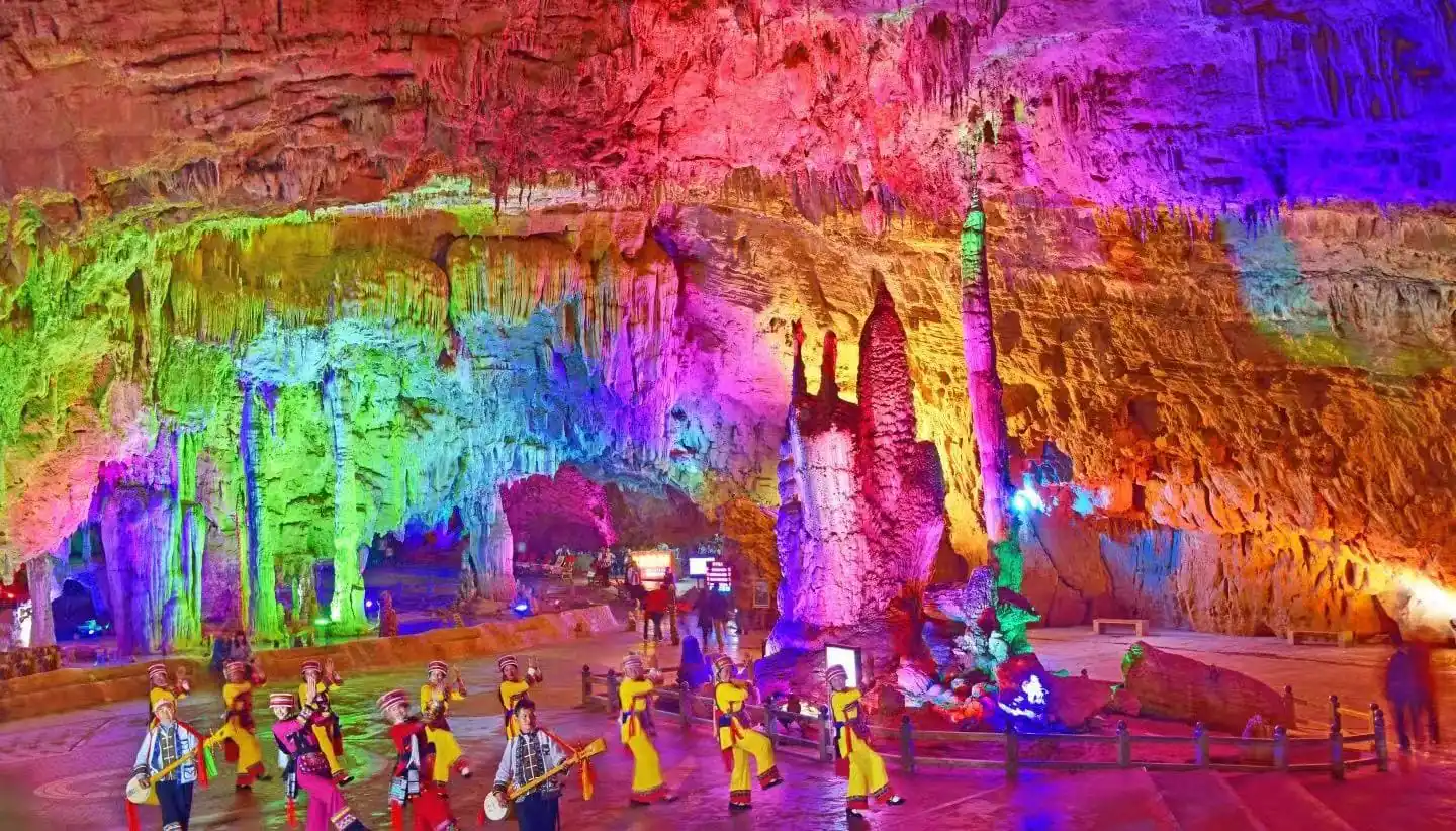 Colorfully lit cave interior with towering rock formations as a group of dancers in yellow traditional outfits perform with musicians in front of stalagmites.Jiuxiang Scenic Area,Kunming,Yunnan ,Chinatravel