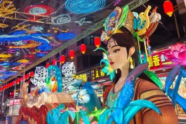 Vibrant Chinese lantern festival scene with a life-size woman statue wearing an ornate headpiece, surrounded by oversized blue leaves and pink lotus flowers beneath a ceiling of colorful neon lanterns.Mojia Subdistrict Night Market,Xining,Qinghai