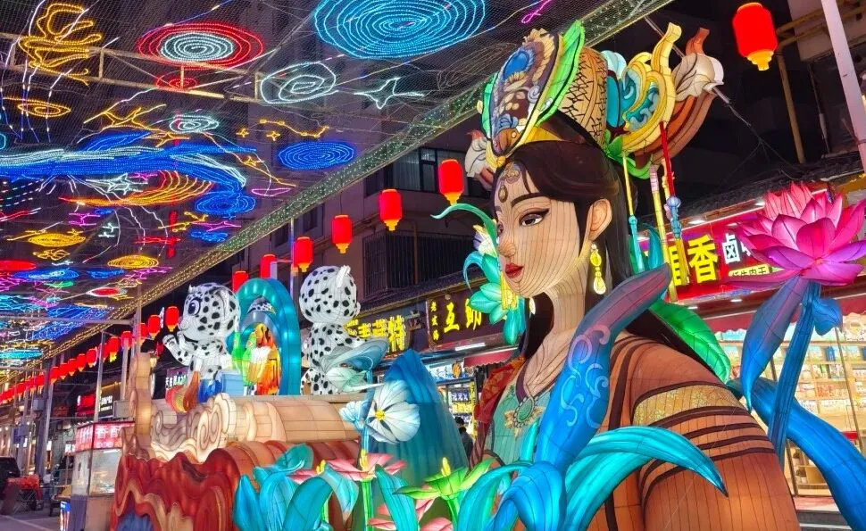 Vibrant Chinese lantern festival scene with a life-size woman statue wearing an ornate headpiece, surrounded by oversized blue leaves and pink lotus flowers beneath a ceiling of colorful neon lanterns.Mojia Subdistrict Night Market,Xining,Qinghai