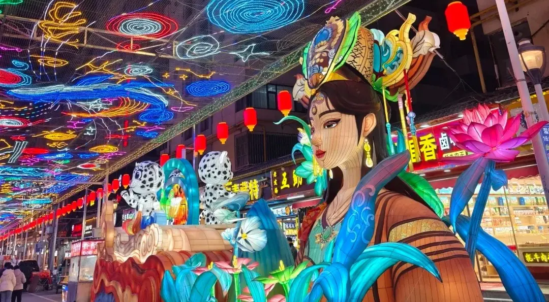 Vibrant Chinese lantern festival scene with a life-size woman statue wearing an ornate headpiece, surrounded by oversized blue leaves and pink lotus flowers beneath a ceiling of colorful neon lanterns.Mojia Subdistrict Night Market,Xining,Qinghai