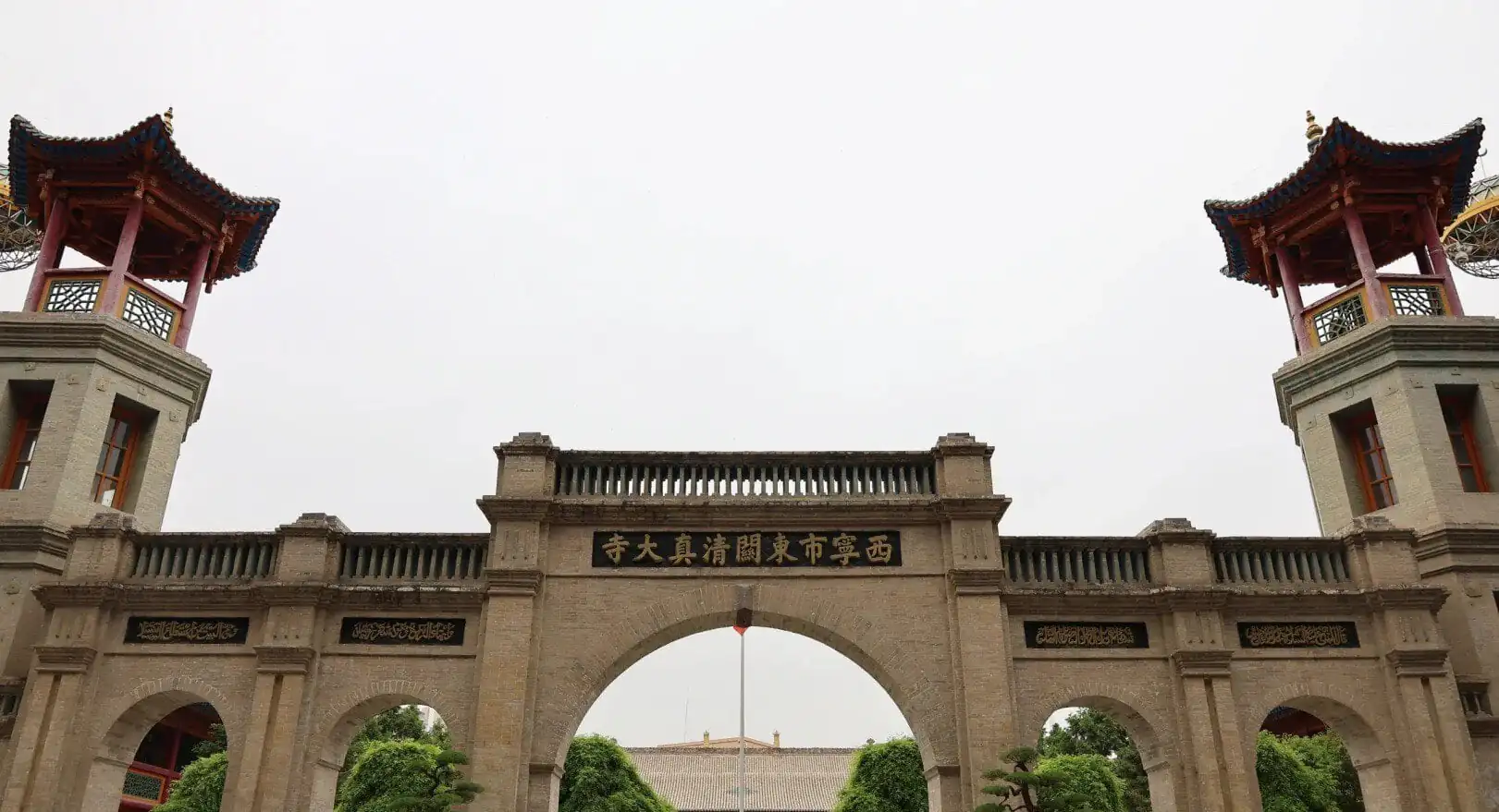 Stone arch gateway in a temple complex, with a central arch flanked by two towers featuring red, pagoda-style roofs and gold Chinese characters on plaques beneath.Dongguan Mosque,Xining,Qinghai