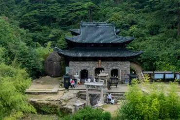 Traditional stone temple with dark tiled, multi-tiered roof nestled in a dense green forest; visitors at the entrance.Sanqing Mountain Scenic Area,Shangrao,Jiangxi,Chinatravel