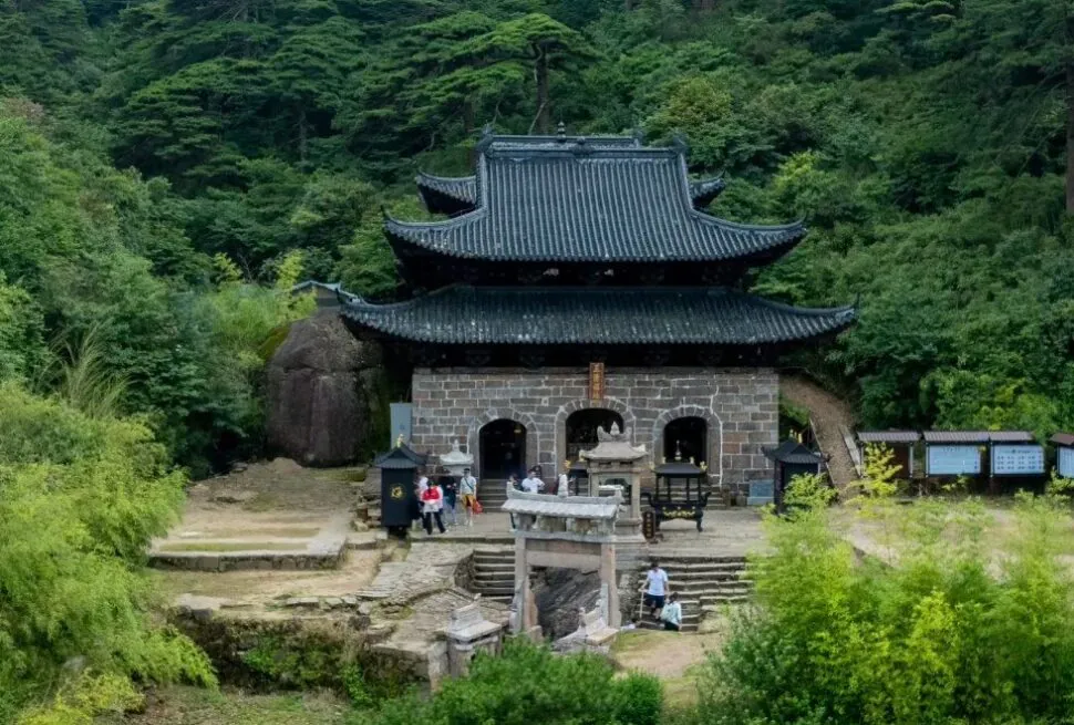 Traditional stone temple with dark tiled, multi-tiered roof nestled in a dense green forest; visitors at the entrance.Sanqing Mountain Scenic Area,Shangrao,Jiangxi,Chinatravel