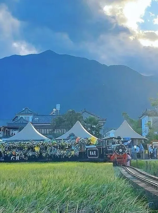 Steam locomotive at a countryside festival on a railway track, with white flower-decorated tents and a crowd, mountains in the distance.Xizhou and Zhoucheng Bai Villages,Dali,Yunnan,Chinatravel
