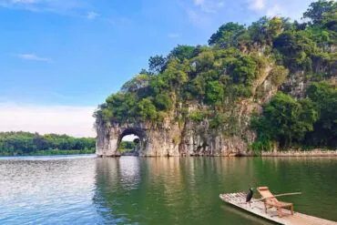 Lush green cliff with a natural rock arch over calm lake, a bamboo raft with chairs docked nearby, and a black bird perched on the raft.Elephant Trunk Hill,Guilin,Guangxi ,Chinatravel
