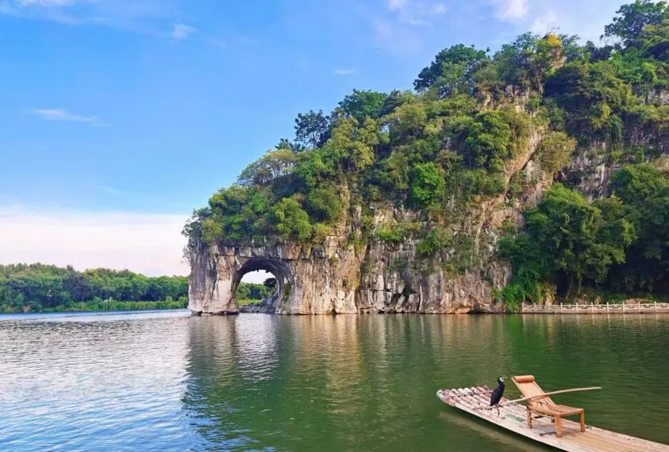 Lush green cliff with a natural rock arch over calm lake, a bamboo raft with chairs docked nearby, and a black bird perched on the raft.Elephant Trunk Hill,Guilin,Guangxi ,Chinatravel