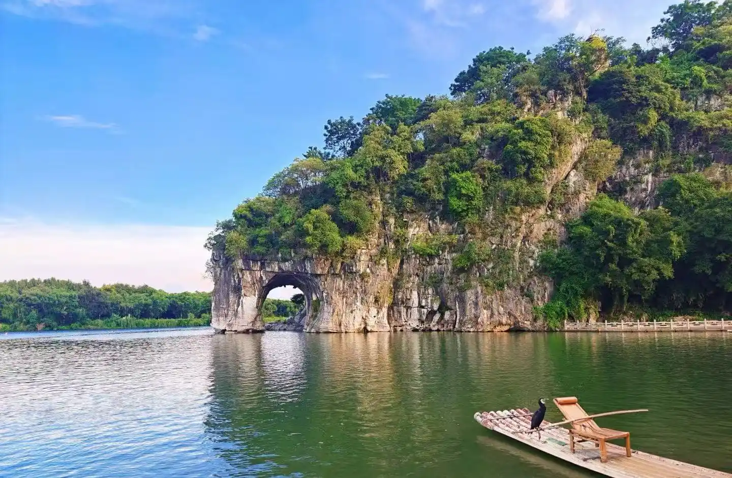 Lush green cliff with a natural rock arch over calm lake, a bamboo raft with chairs docked nearby, and a black bird perched on the raft.Elephant Trunk Hill,Guilin,Guangxi ,Chinatravel