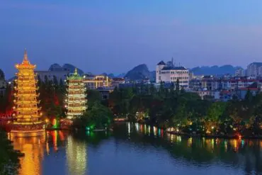 Two illuminated tiered pagodas beside a calm lake, reflections glowing gold and green with a cityscape in the background.Sun and Moon Twin Pagodas,Guilin,Guangxi ,Chinatravel