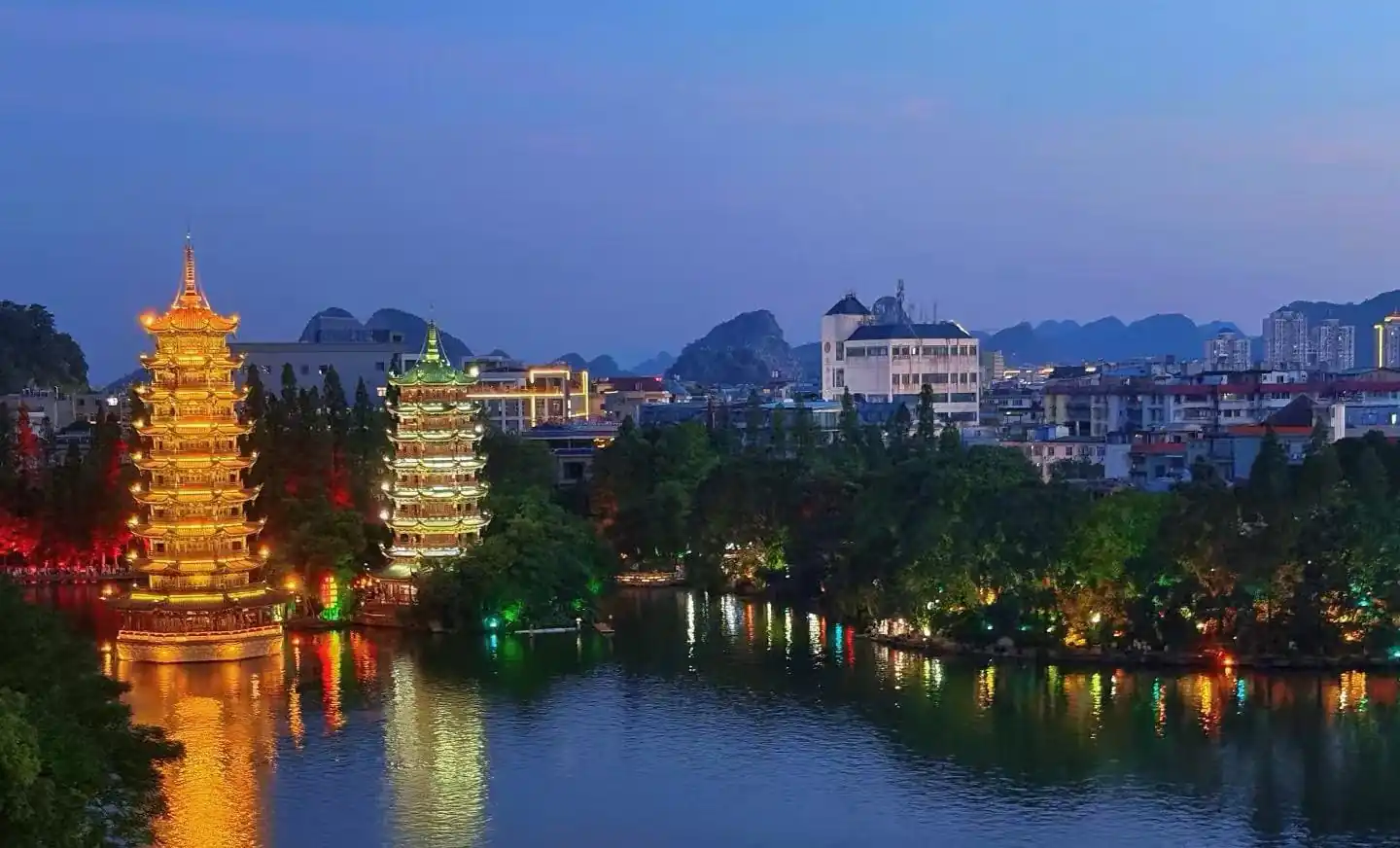 Two illuminated tiered pagodas beside a calm lake, reflections glowing gold and green with a cityscape in the background.Sun and Moon Twin Pagodas,Guilin,Guangxi ,Chinatravel