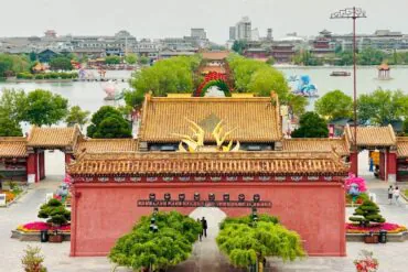 Dragon Pavilion Park & Park,Kaifeng,Henan,Chinatravel Red-walled Chinese temple entrance with golden tiled roofs and a central arch, leading to a tree-lined courtyard and pond in the distance.