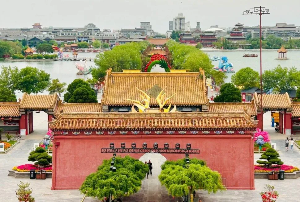 Dragon Pavilion Park & Park,Kaifeng,Henan,Chinatravel Red-walled Chinese temple entrance with golden tiled roofs and a central arch, leading to a tree-lined courtyard and pond in the distance.