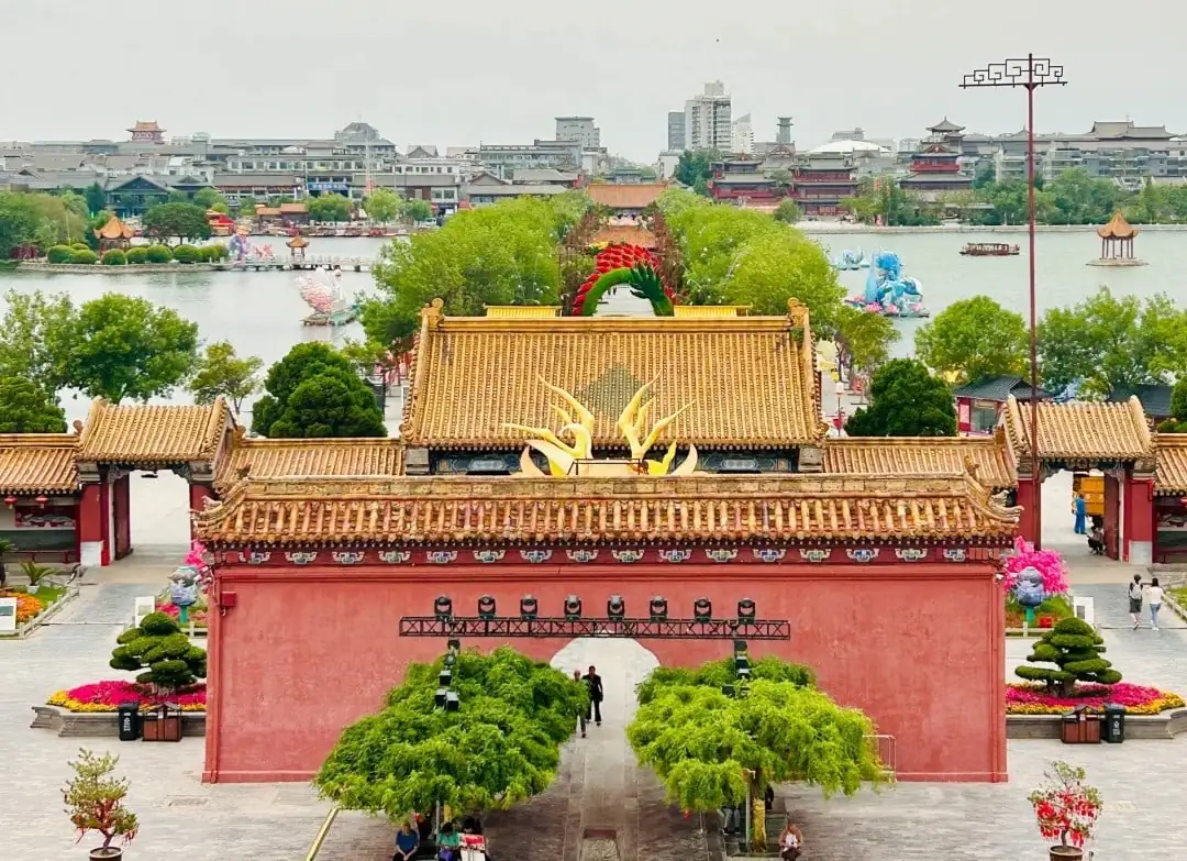 Dragon Pavilion Park & Park,Kaifeng,Henan,Chinatravel Red-walled Chinese temple entrance with golden tiled roofs and a central arch, leading to a tree-lined courtyard and pond in the distance.