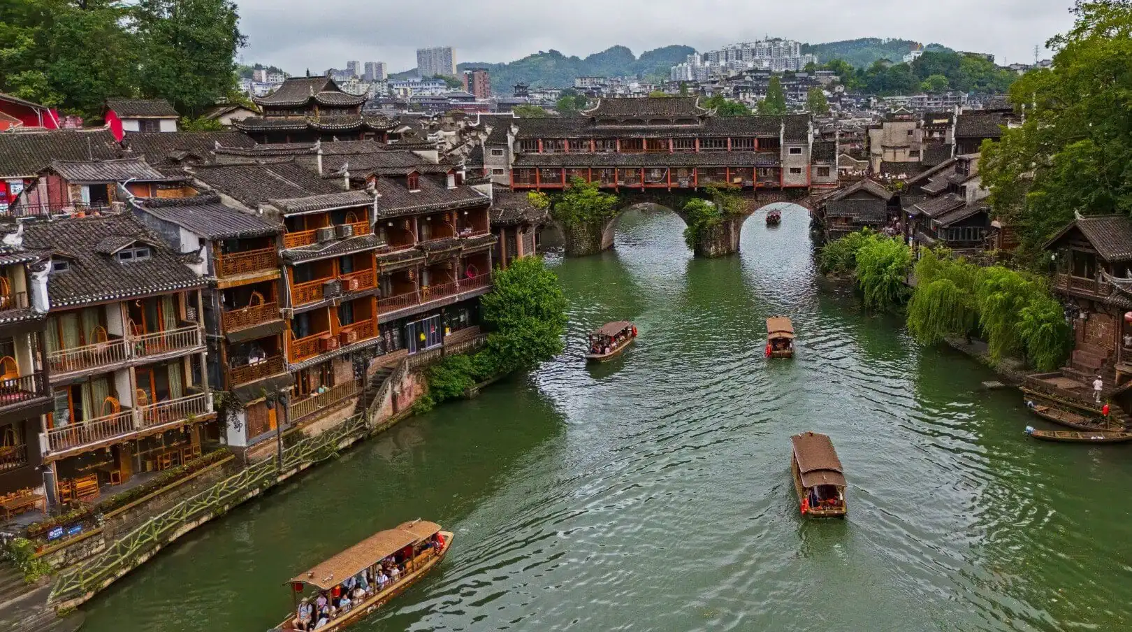 Fenghuang Ancient Town,Fenghuang,Hunan,Chinaexplorertour Ancient riverside town with wooden shophouses lining a green canal and a stone arch bridge, boats gliding on the water.