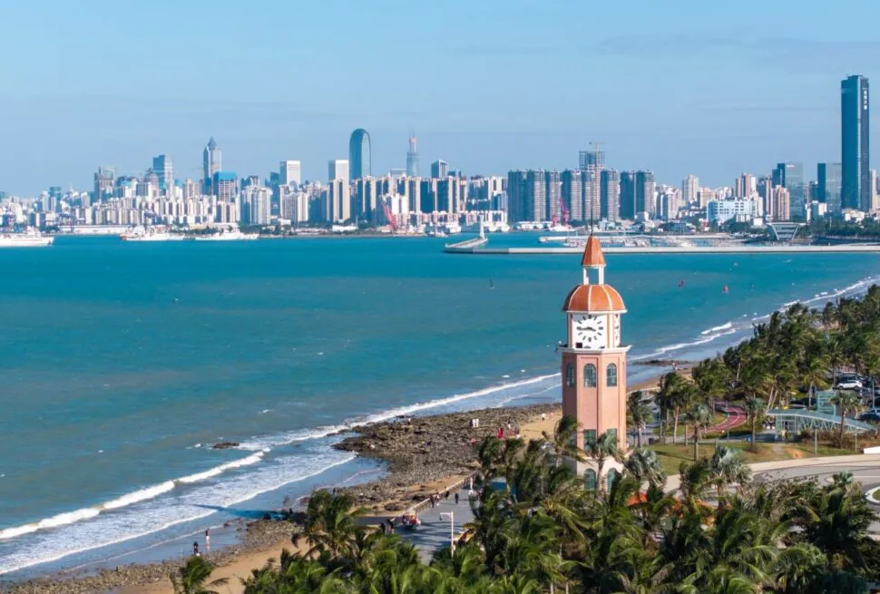 Holiday Beach,Haikou,Hainan,Chinaexplorertour Clock tower with orange dome near a palm-lined promenade overlooking a blue sea and city skyline across the water.