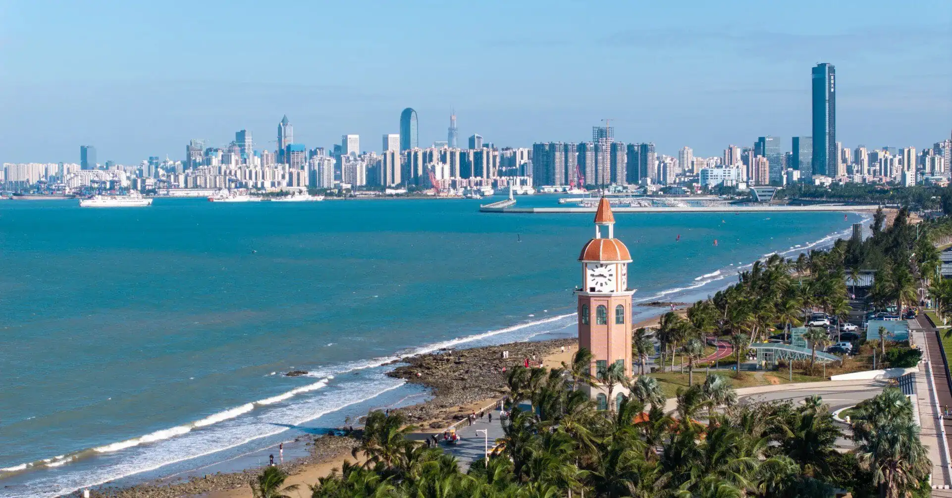 Holiday Beach,Haikou,Hainan,Chinaexplorertour Clock tower with orange dome near a palm-lined promenade overlooking a blue sea and city skyline across the water.
