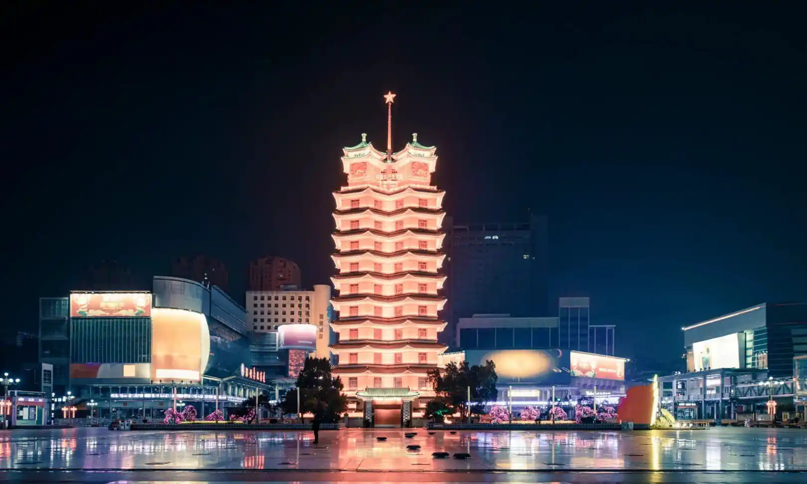 Erqi Memorial Tower,Zhengzhou,Henan,Chinaexplorertour Night view of a lit multi‑tiered pagoda tower in a city square, its warm glow reflected on the wet pavement.
