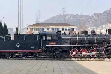 Dalian Modern Museum,Dalian,Liaoning,Chinatravel Historic steam locomotive on display in a public square; black body with red wheels and white piping, mountainous backdrop.